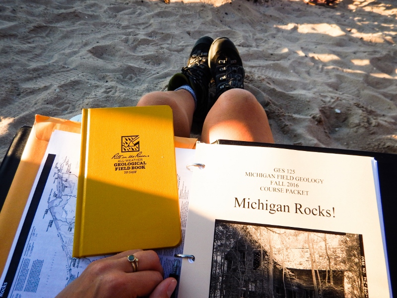 Michigan Fied Geology binder and field book on a student's lap with legs outstretched and hiking boots resting in beach sand