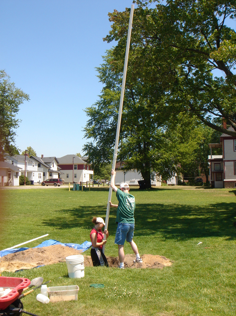 students guiding a 20 foot PVC pipe water well into a hole on a campus lawn