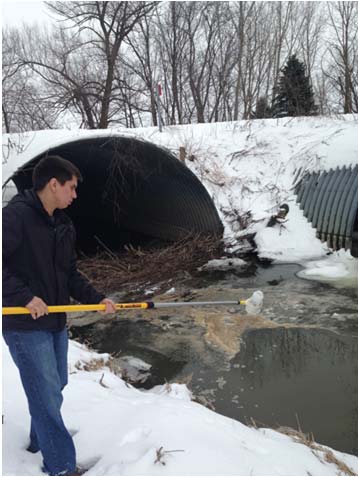 student holds a pole with a sampling bottle on the end over a stream by a snow-covered culvert