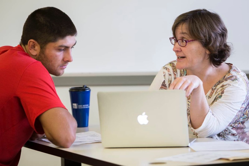 faculty member advising a student. A computer sits on the table between them.