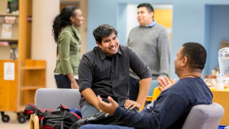 A faculty member and student talking in the library. Two other people are standing up having a conversation in the background.